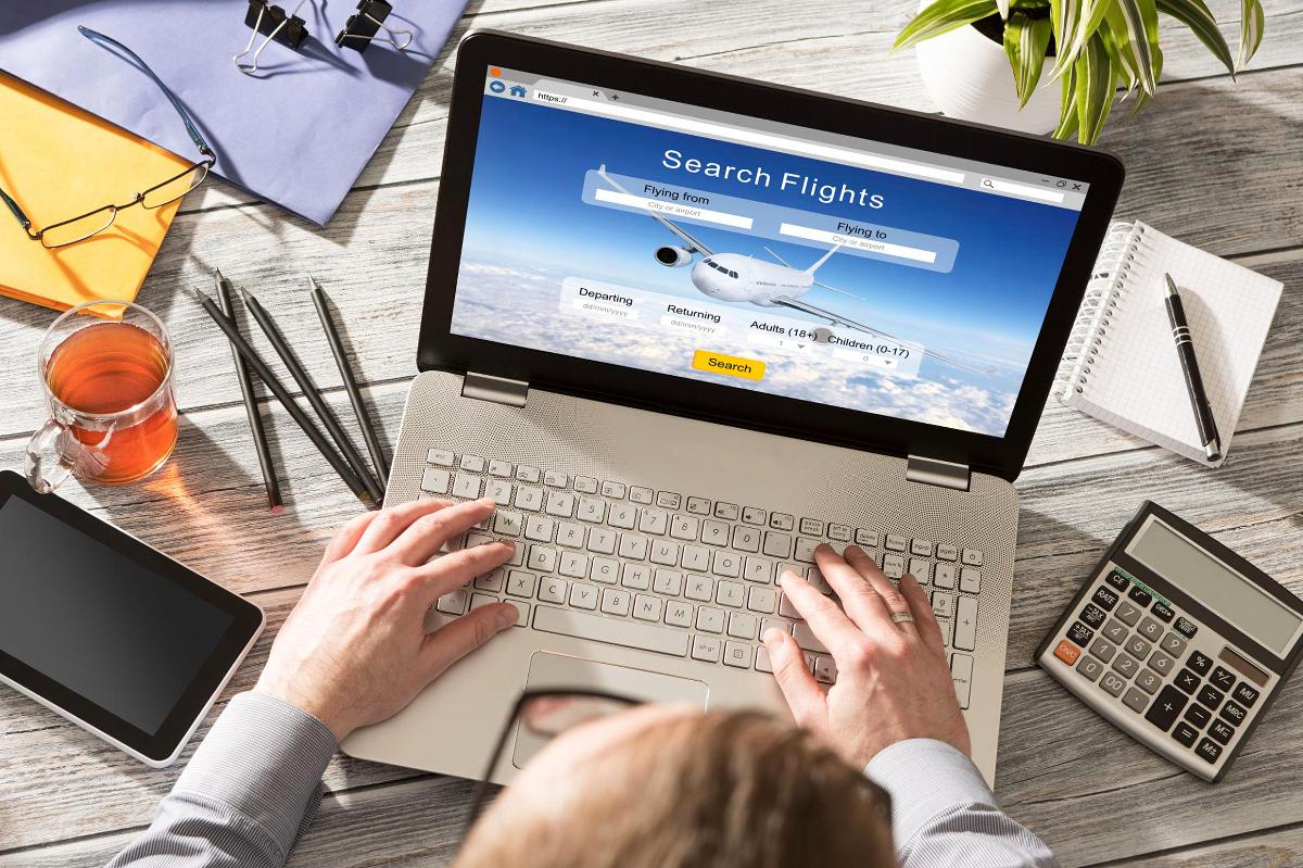 Hände tippen auf einem Laptop, der eine Buchungsseite 'Flüge suchen' mit Flugzeuggestaltung anzeigt, neben einem Notizbuch, einem Taschenrechner und einer Tasse Tee.