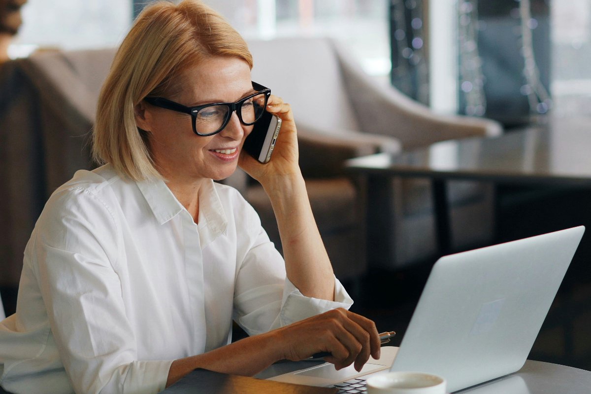 Frau mittleren Alters mit Brille, lachend am Telefon und an einem Laptop in einem Café arbeitend — Die eigentliche Prüfung beginnt, nachdem Sie bezahlt haben