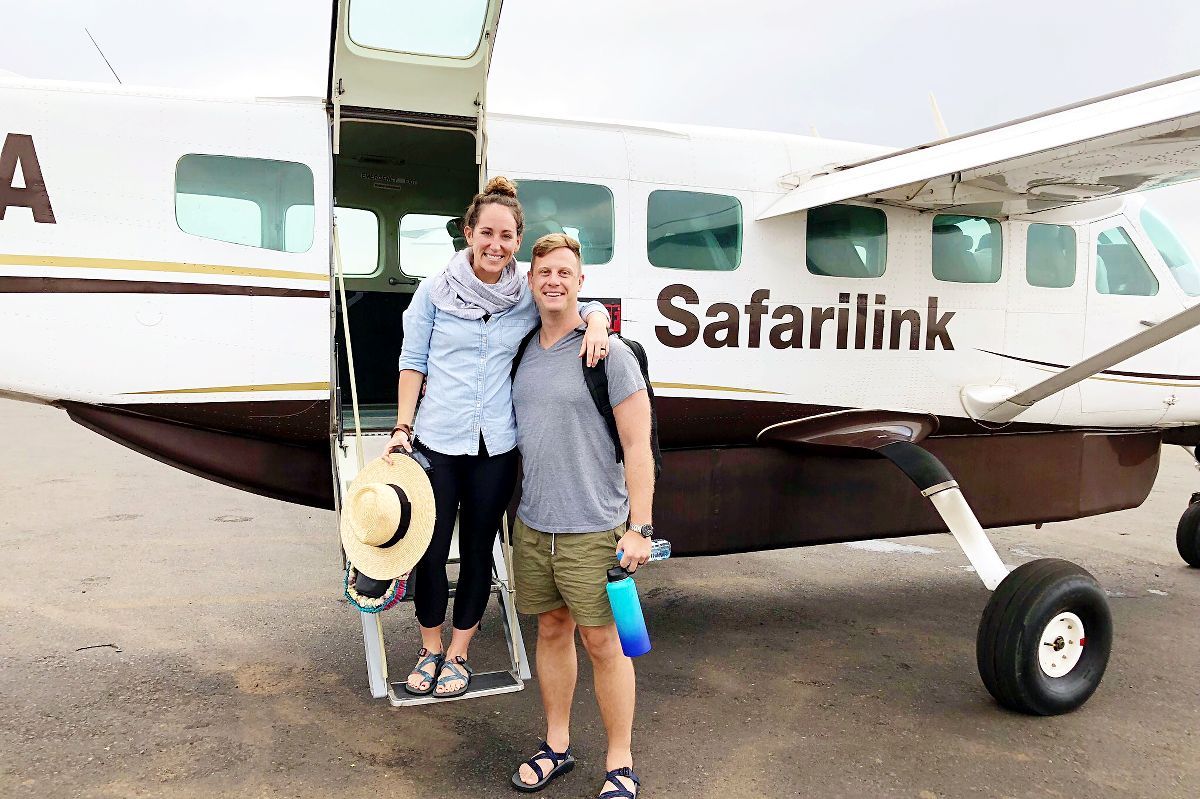 Paar lächelnd auf der Einstiegsleiter einer Safarilink-Turboprop-Maschine, Frau hält einen Strohhut, Mann eine Wasserflasche — Wählen Sie Ihren Safari-Reisestil