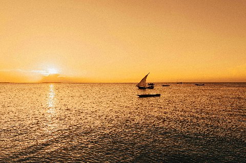Traditional dhow sailing at sunset off Zanzibar City, orange sky and silhouetted boats on calm seas