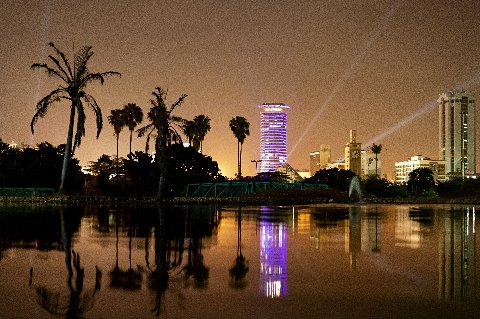 Nighttime Nairobi scene with an illuminated circular tower and palm trees mirrored in still water