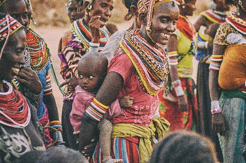 Women wearing colorful beaded necklaces and bracelets smiling and chatting during a village gathering