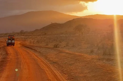 Un veicolo safari scoperto percorre una pista di terra rossa verso un tramonto infuocato sulle pianure di Tsavo